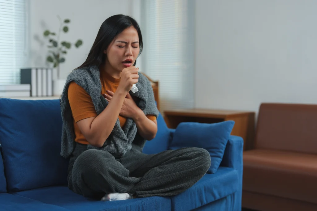 Woman sitting on a couch holding her chest and coughing, showing symptoms of a chest infection and breathing difficulty at home in Katy, TX.