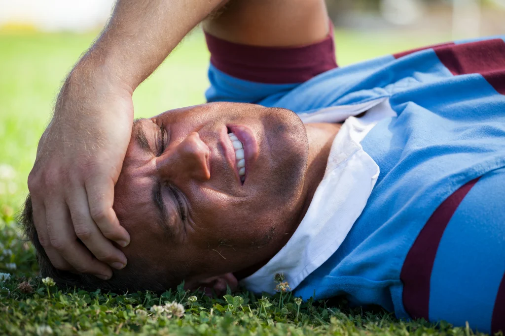 Man lying on the grass holding his head in pain after a sports injury, illustrating concussion signs that may require emergency care in Katy.